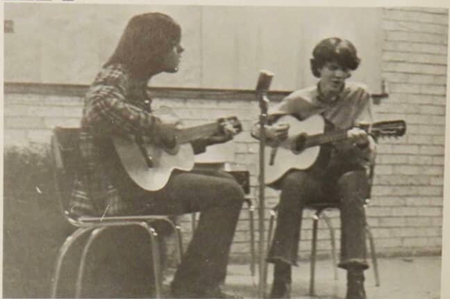 Black & white photo of two people sit in chairs, playing guitar.
